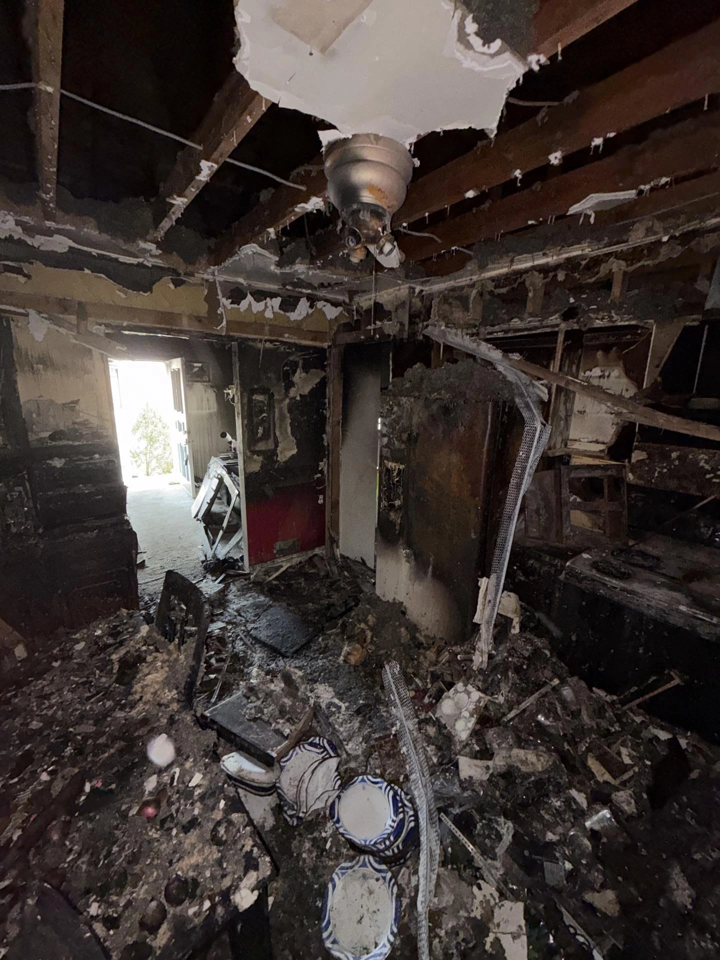 Interior of a fire-damaged house showing charred walls, debris on the floor, and a damaged ceiling fan, illustrating the aftermath of smoke and soot damage requiring professional fire restoration services.