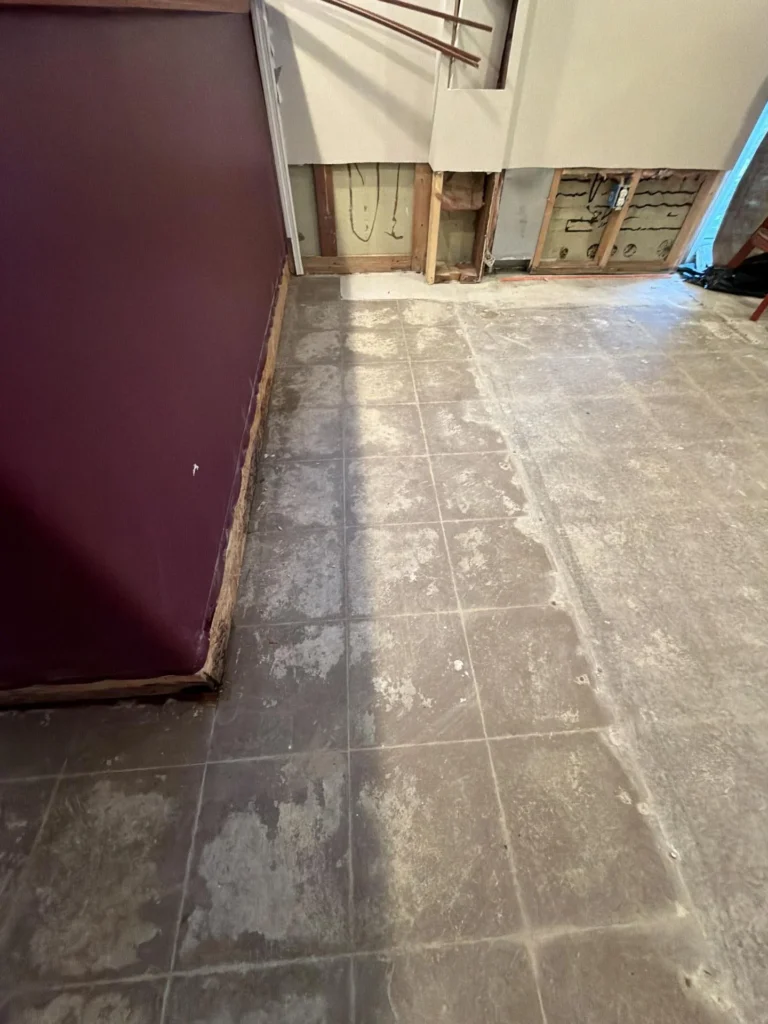 Wide view of basement bar area with dark burgundy bar on left side, dirty gray tile floor showing water damage staining, exposed wall framing with green insulation board and wiring visible through multiple drywall cutouts, copper pipe running along wall in background