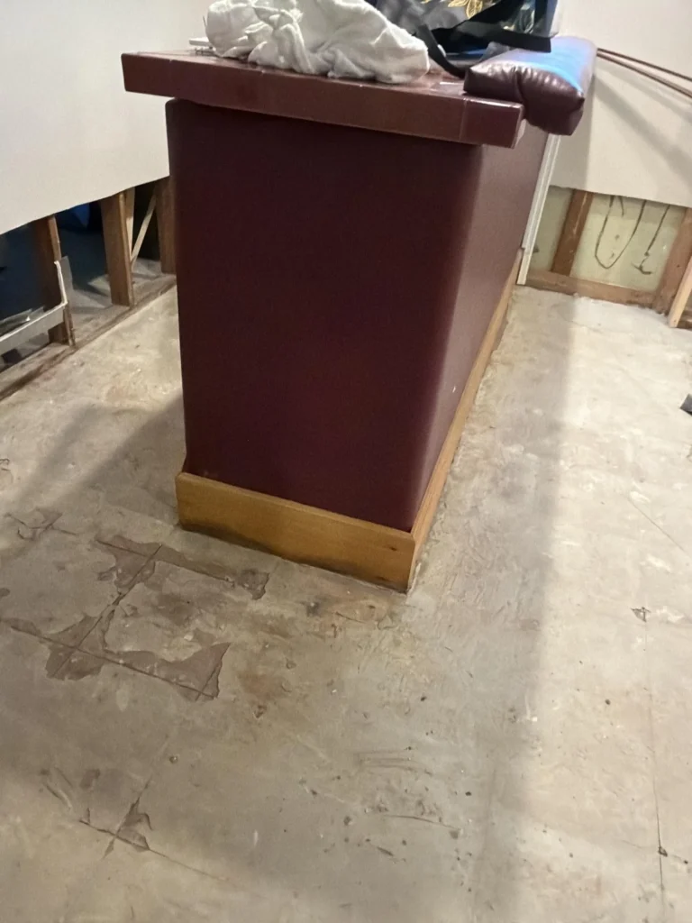 Back side of dark burgundy basement bar unit on stripped concrete subfloor, exposed wall framing with HVAC ductwork visible behind, misc items on top of bar, blue object in background