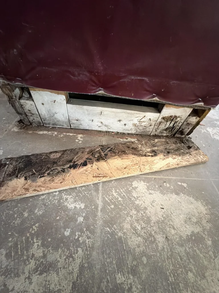 Extreme close-up of bottom section of basement bar cabinet showing severely mold-blackened and water-damaged OSB board and wood framing at floor level, warped vinyl skin peeling away from base, tile floor with dust and debris