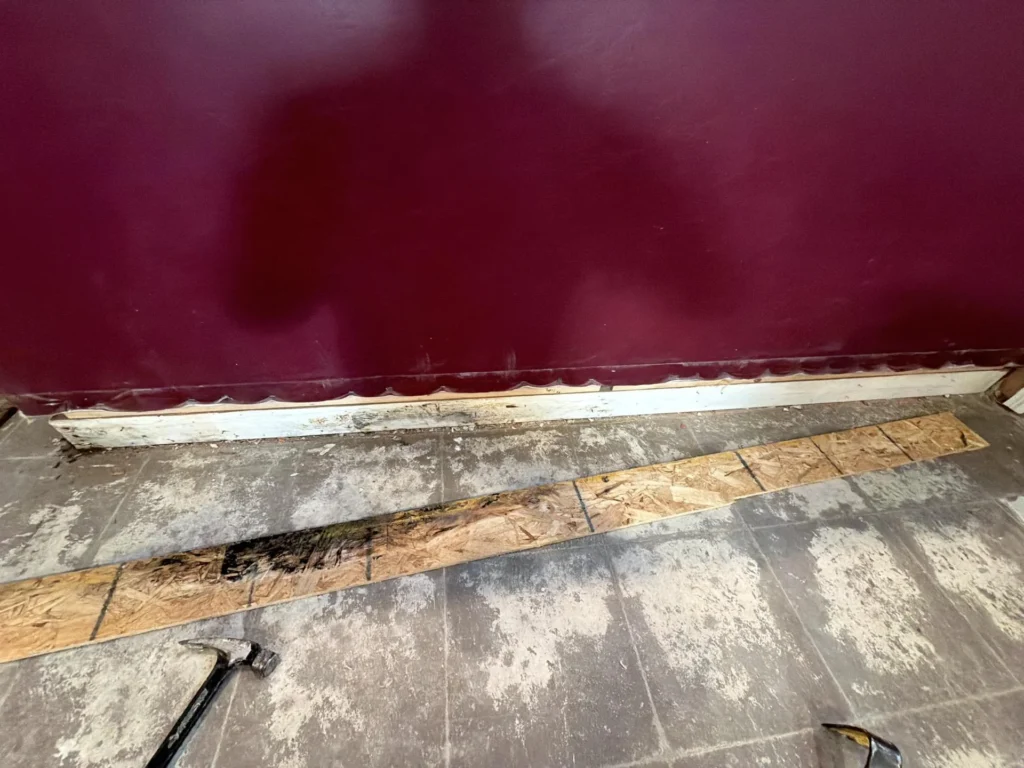 Close-up of bottom of dark burgundy basement bar showing removed baseboard trim exposing water-damaged and mold-stained OSB sheathing and base plate, two dark pry bars on tile floor, demolition in progress