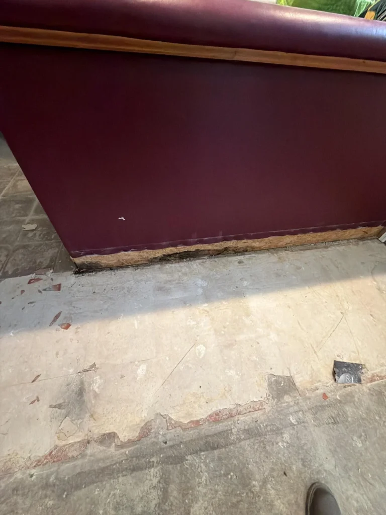 Close-up of dark burgundy basement bar base showing severely water-damaged and rotted OSB board with black mold at the bottom, peeling flooring debris on concrete subfloor nearby
