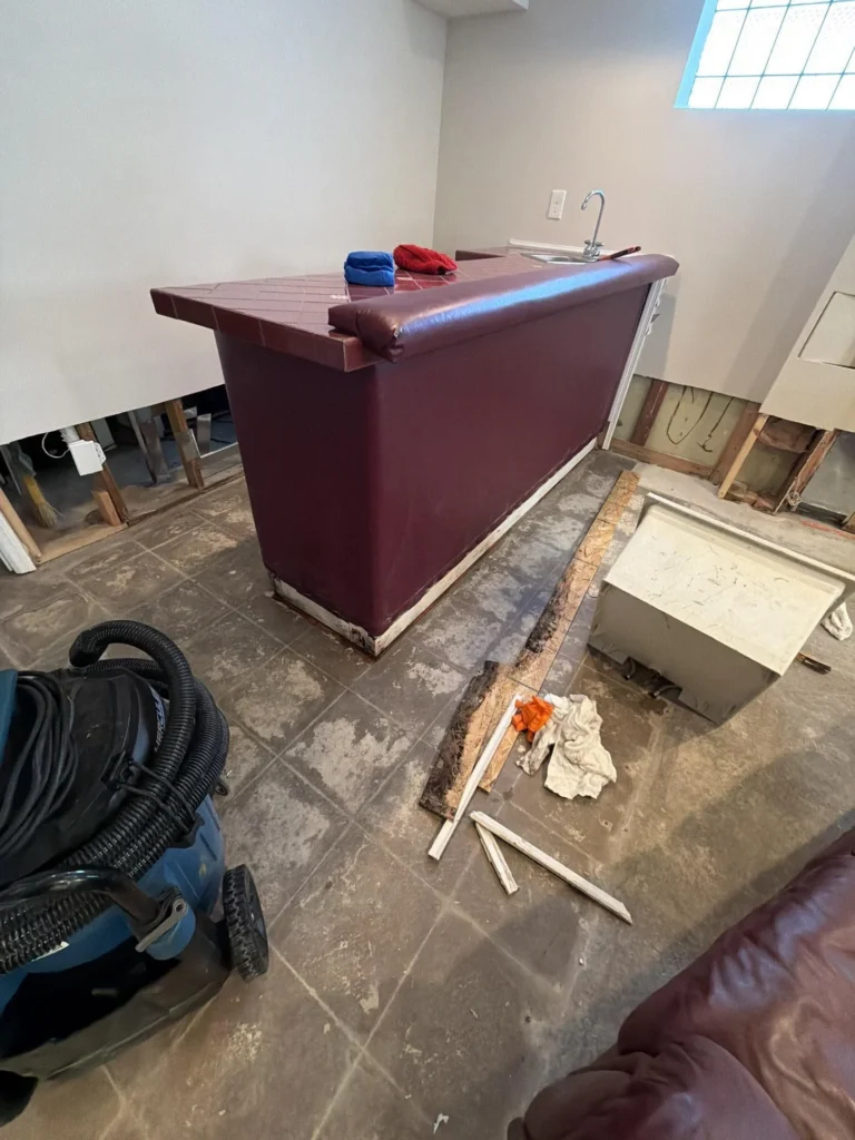 Full view of freestanding basement wet bar with dark burgundy laminate sides and tiled countertop with stainless sink, blue wet-dry vacuum on left, damaged OSB subfloor boards removed and lying on tile, exposed wall framing with HVAC duct in background, glass block window above
