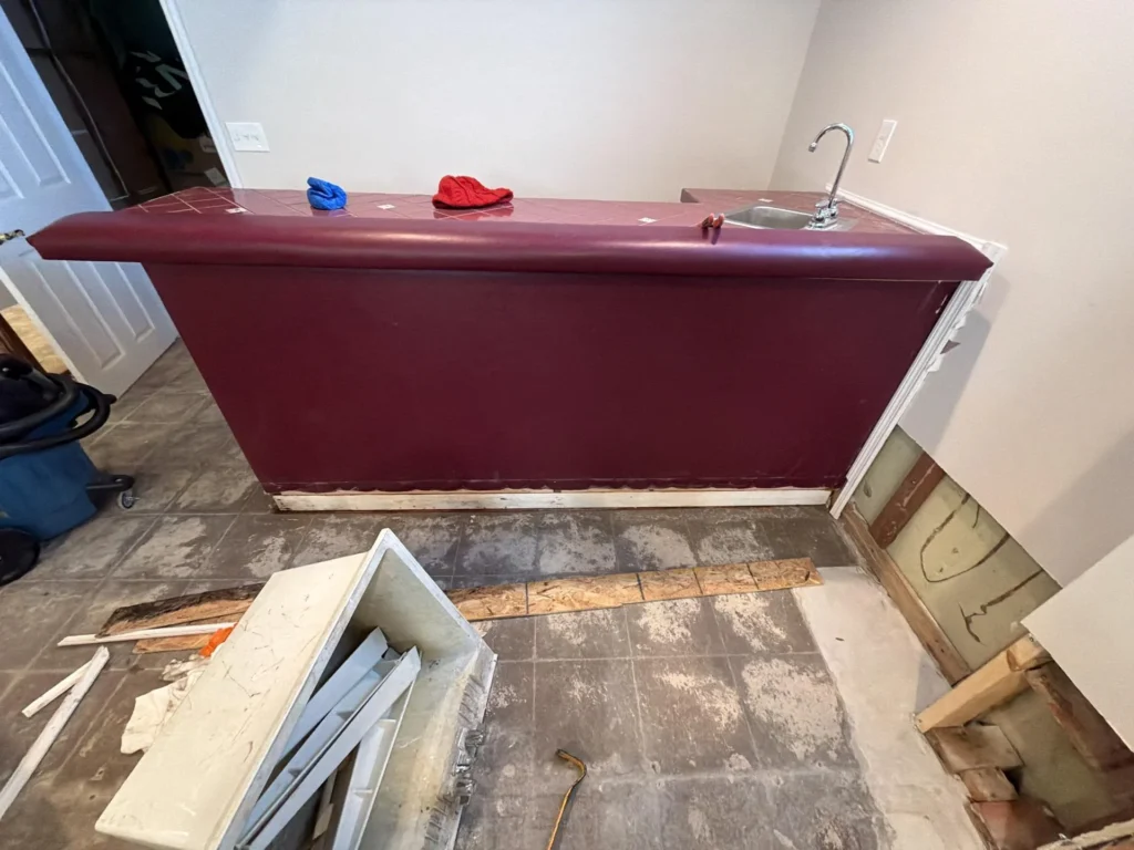 Basement wet bar with dark burgundy laminate front panel and tiled top surface with stainless steel sink and chrome gooseneck faucet, removed HVAC register box on floor in foreground, exposed wall framing on right, demolition debris scattered on tile