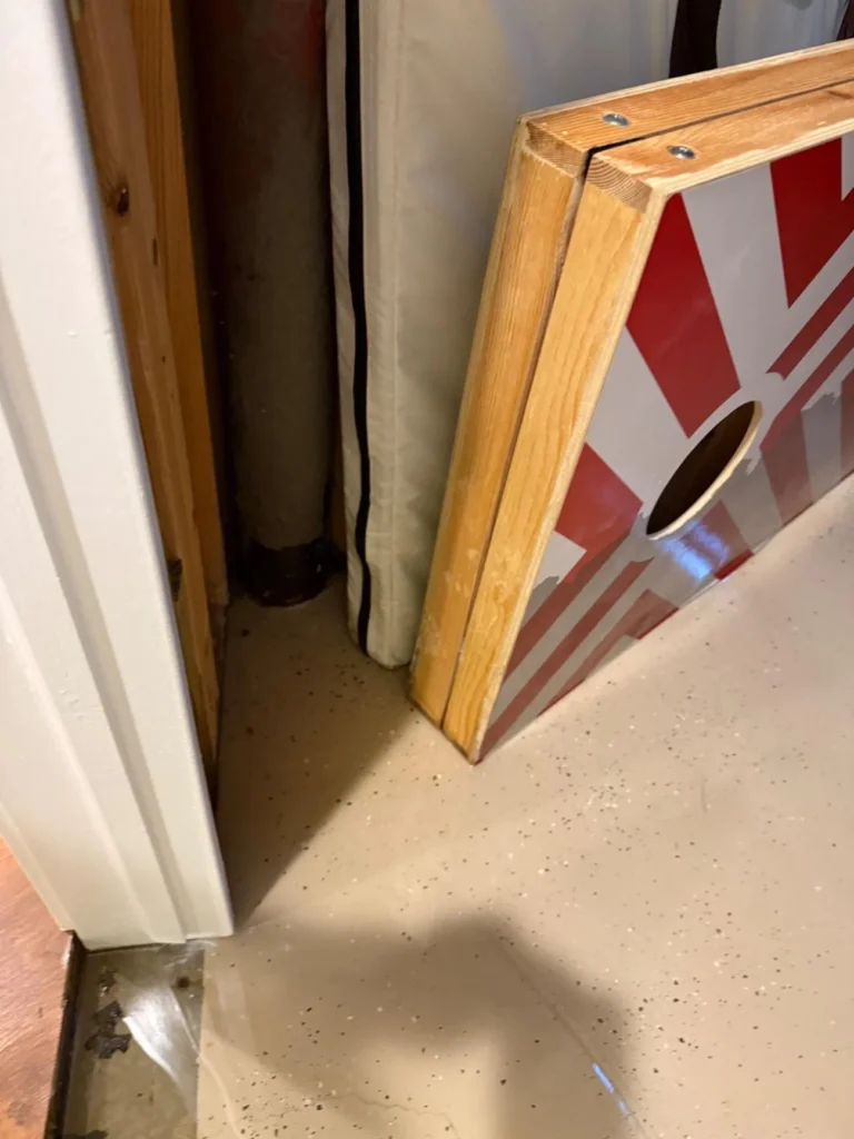 Basement doorway showing water intrusion at threshold with cornhole game board leaning against wall and exposed drain pipe visible