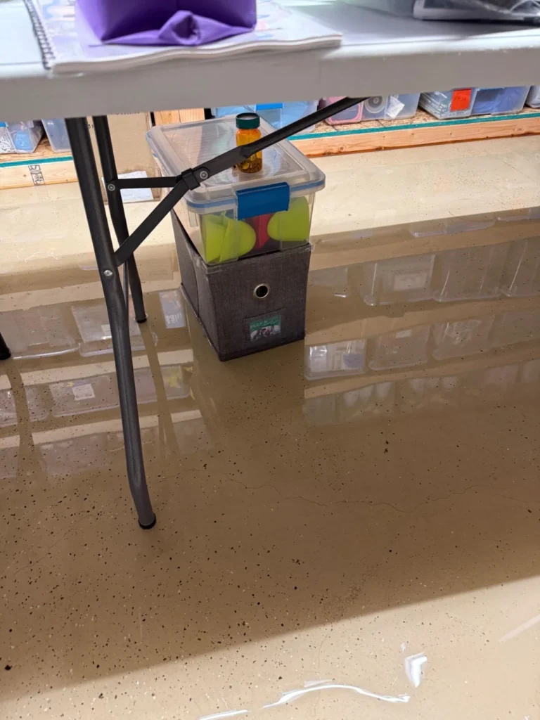 Close-up of standing flood water under folding table in basement, storage bin and fabric box sitting in shallow water on epoxy floor