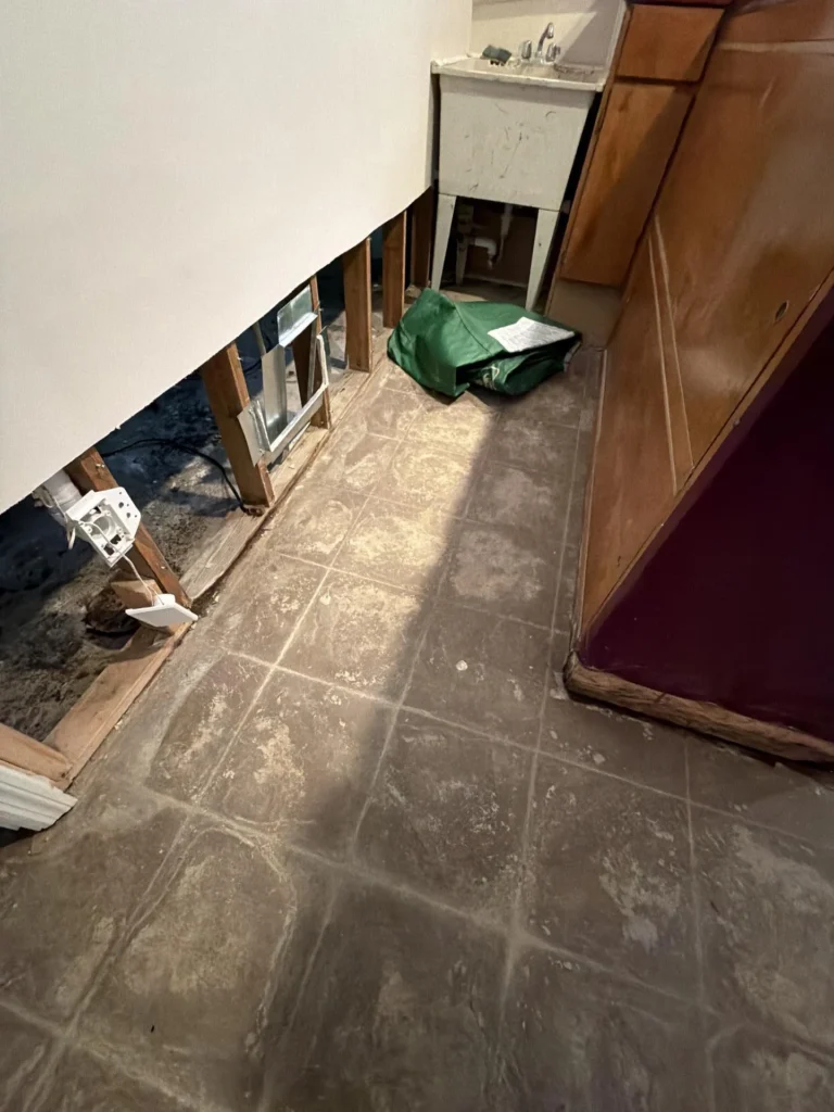 Basement kitchenette area under water damage restoration showing exposed wall framing with electrical box, dirty tile floor with adhesive residue, wood cabinet panels on right, utility sink visible in background, and green bag on floor