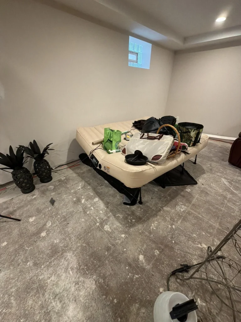 Finished basement rec room with white walls and recessed lighting showing stripped concrete subfloor during renovation, air mattress with belongings in center, decorative pineapple figures on left, miscellaneous items scattered throughout