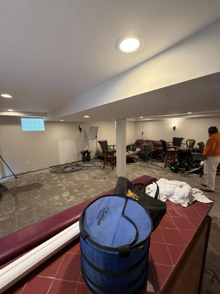 Basement rec room viewed from behind burgundy bar unit showing stripped concrete subfloor, person in orange jacket on right, bar stools and furniture piled in background, blue collapsible bag on bar top, recessed lighting illuminating white walls