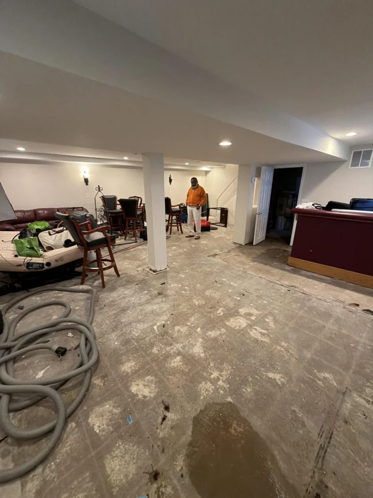 Wide angle view of finished basement rec room during water damage remediation, white walls with recessed lighting and center support column, stripped concrete subfloor, worker in orange shirt standing in background, bar unit and bar stools visible, vacuum hose on floor
