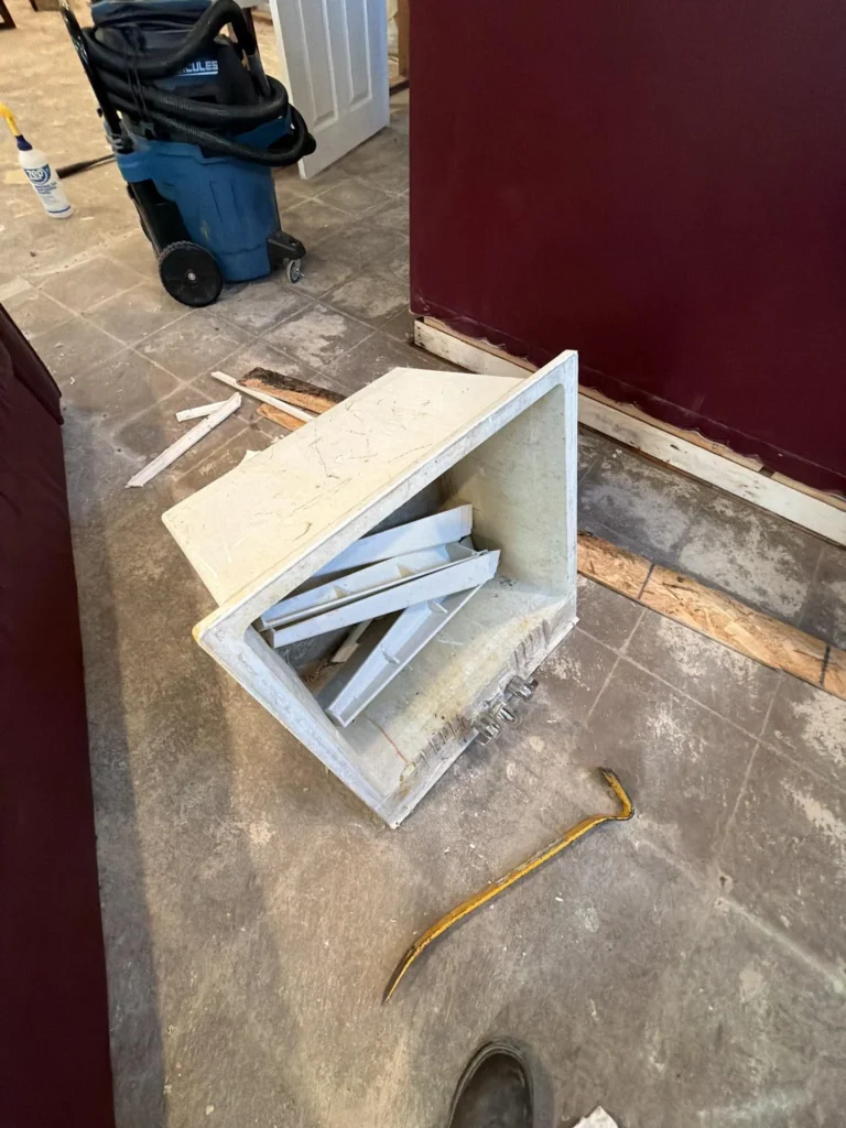 Removed rectangular HVAC floor register box lying open on dirty basement tile floor revealing inner louver assembly, crowbar and pry bar nearby, blue wet-dry vacuum in background, dark burgundy bar panel visible