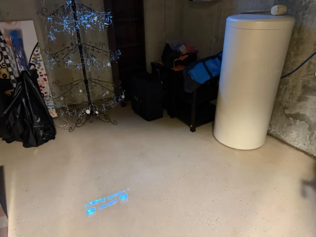 Basement corner with cylindrical water softener or filter tank, decorative iron rack with crystal decorations, and storage items on dry epoxy floor