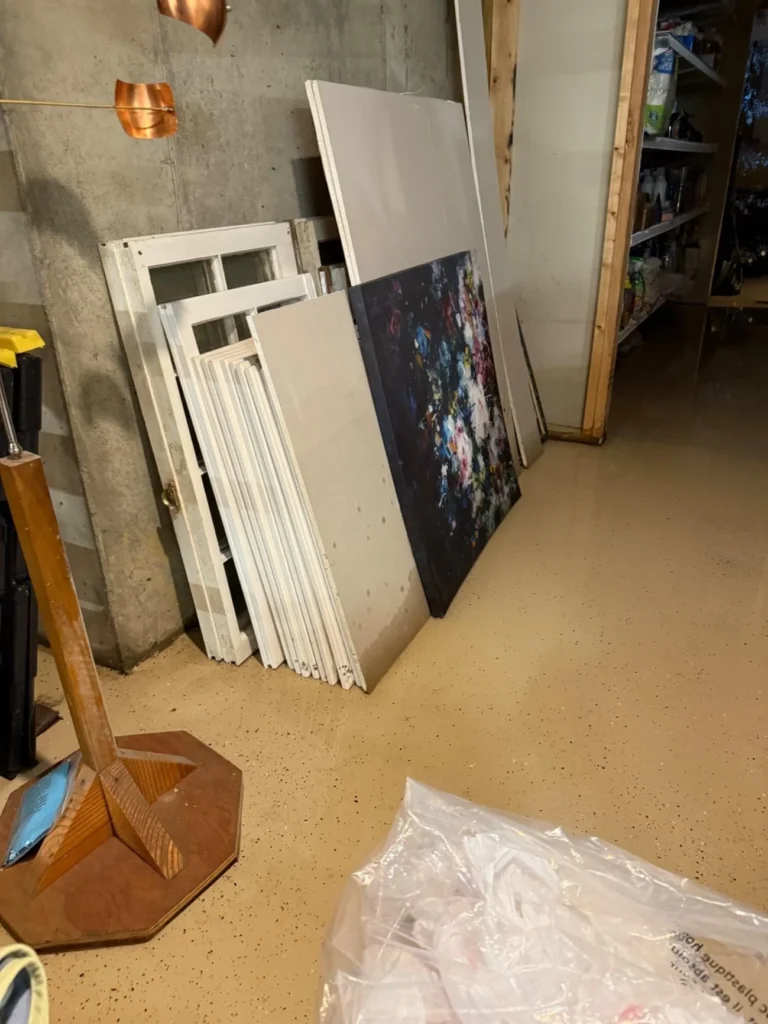 Basement storage area with door frames, artwork, and panels leaning against concrete wall on epoxy floor after water damage event