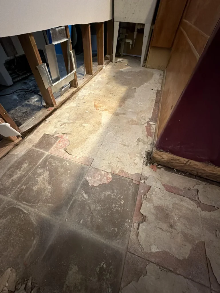 Basement floor view showing gray tile with large sections of peeling red flooring underlayer, exposed wood wall framing with HVAC duct visible, dark burgundy bar panel on right, demolition debris throughout