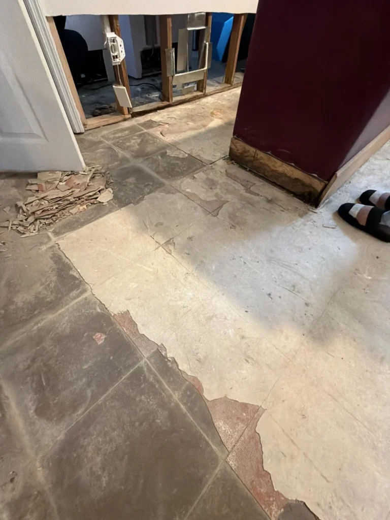 Basement floor during water damage remediation showing gray tile with red underlayer peeling in large sections, exposed wall framing in background, white door on left, burgundy bar panel on right, debris and sandals on floor
