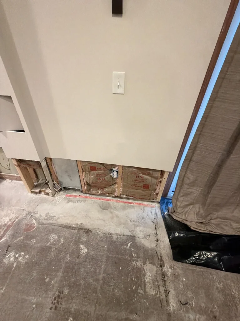 Basement wall with lower drywall section cut out exposing R-13 kraft-faced batt insulation, metal HVAC duct, and wood framing, stripped concrete subfloor with red flooring adhesive residue, black plastic sheeting on right side