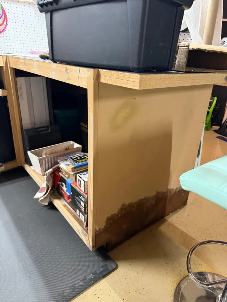 Basement workbench showing water-damaged and swollen particle board panel with dark water stain at base, storage bins and tools on shelving