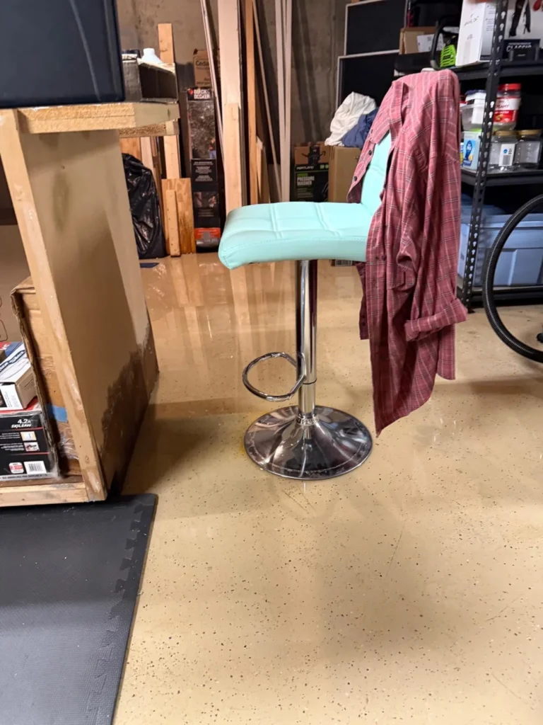 Basement workshop with standing water on epoxy floor, teal bar stool in foreground and water-stained wooden workbench visible in background