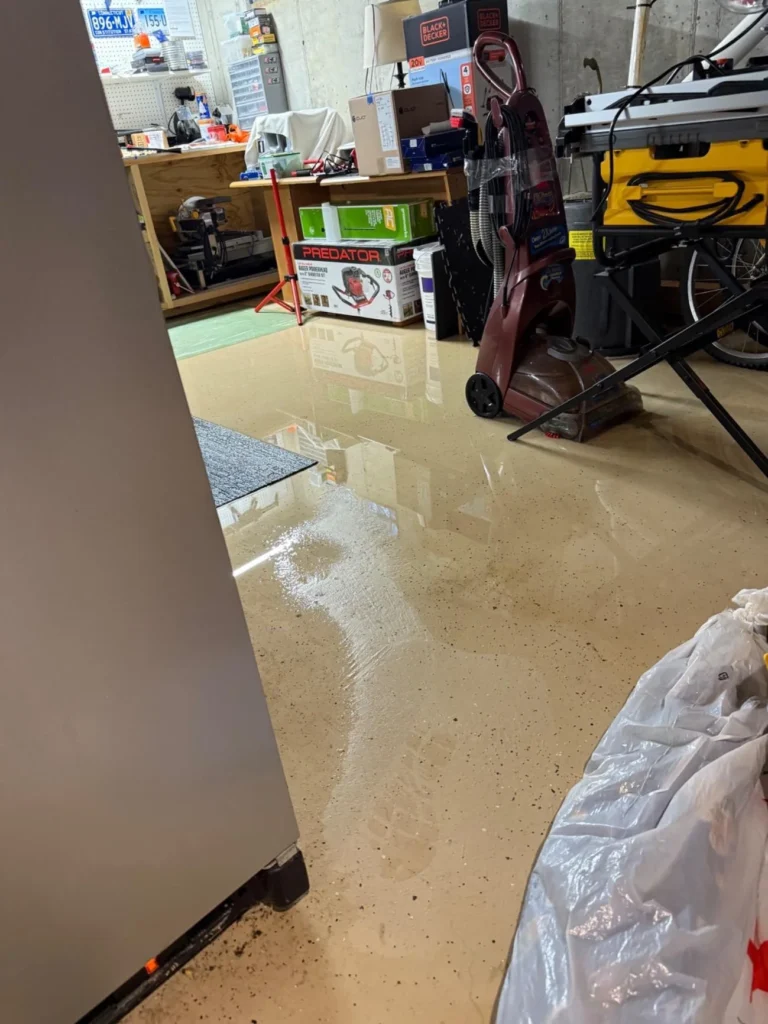 Flooded basement workshop with standing water on epoxy floor, power tools, vacuum, and equipment on shelving in background