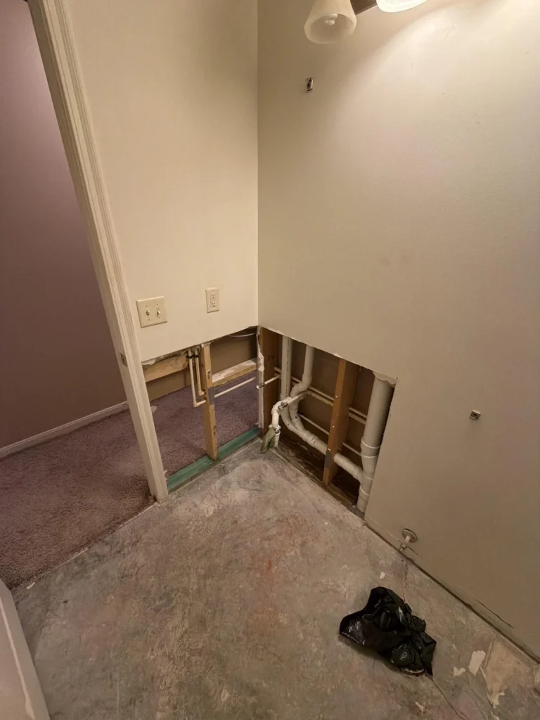 Corner of bathroom showing large drywall cutout revealing PVC drain pipes and wood framing, bare concrete subfloor, overhead vanity light, and open doorway to carpeted hallway