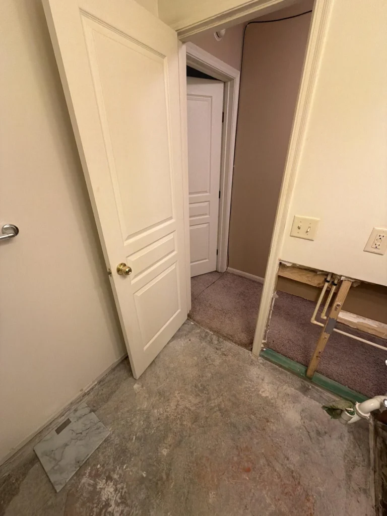 View from bathroom doorway showing exposed PVC and copper plumbing pipes through open drywall, bare concrete subfloor, open door leading to carpeted hallway with brown walls