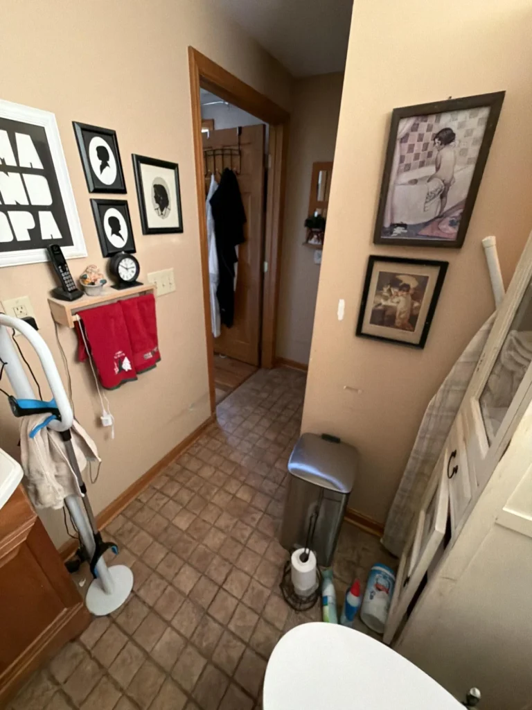 Bathroom interior showing tile floor, framed silhouette portraits and vintage wall art, a towel bar, trash can, and open doorway leading to a bedroom closet area