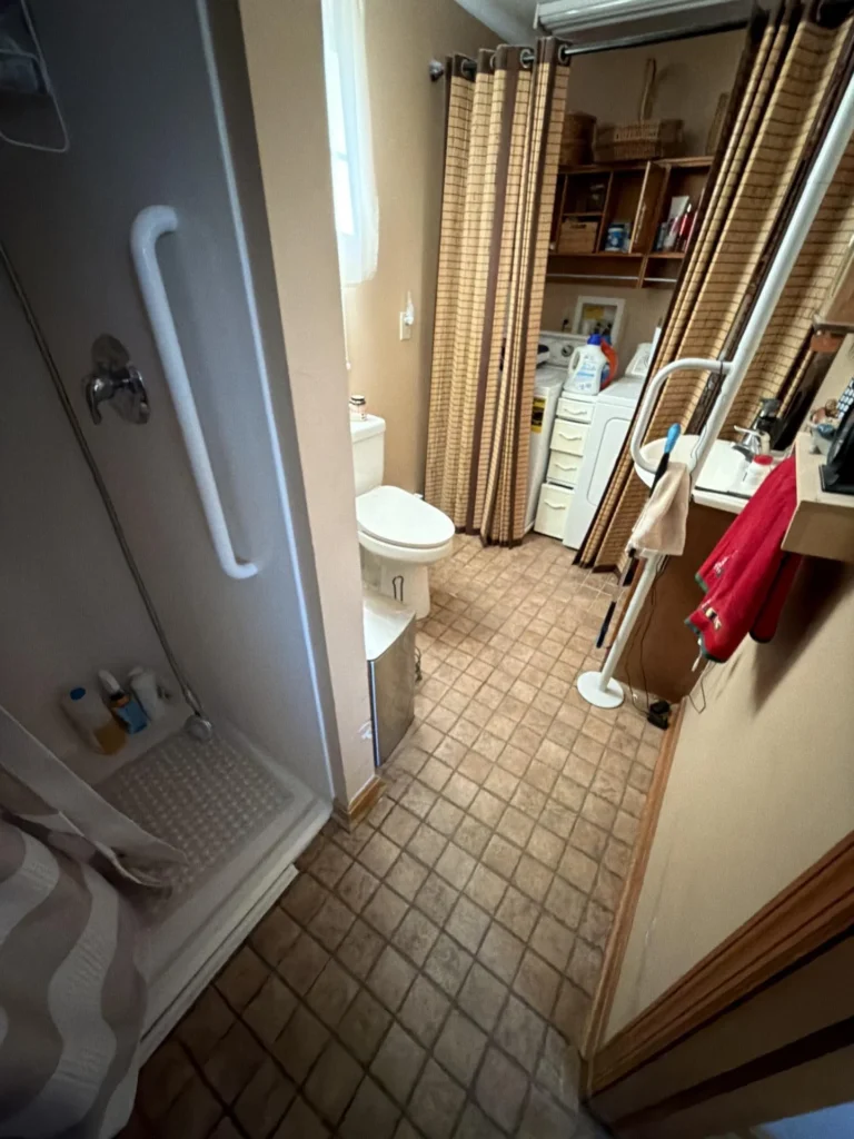 Overview of a narrow bathroom and laundry room combination with tile floor, walk-in shower with grab bar, toilet, pedestal sink, washer and dryer, and open shelving before renovation