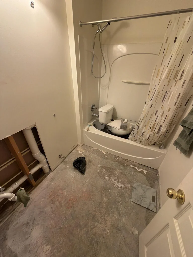 Wide overhead view of bathroom under renovation with white tub-shower combo, handheld showerhead, exposed plumbing pipes through open drywall on left wall, stripped concrete floor, and white door
