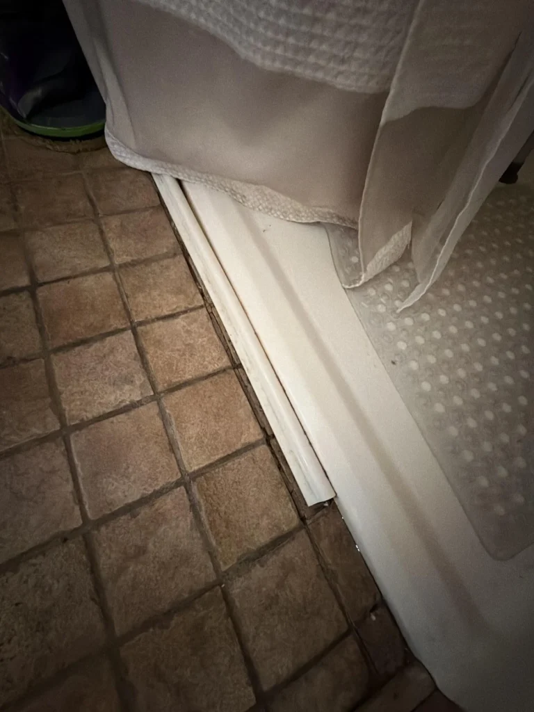 Close-up of a bathroom shower threshold showing deteriorated caulk or seal between the tile floor and shower pan base, indicating water damage or wear needing repair