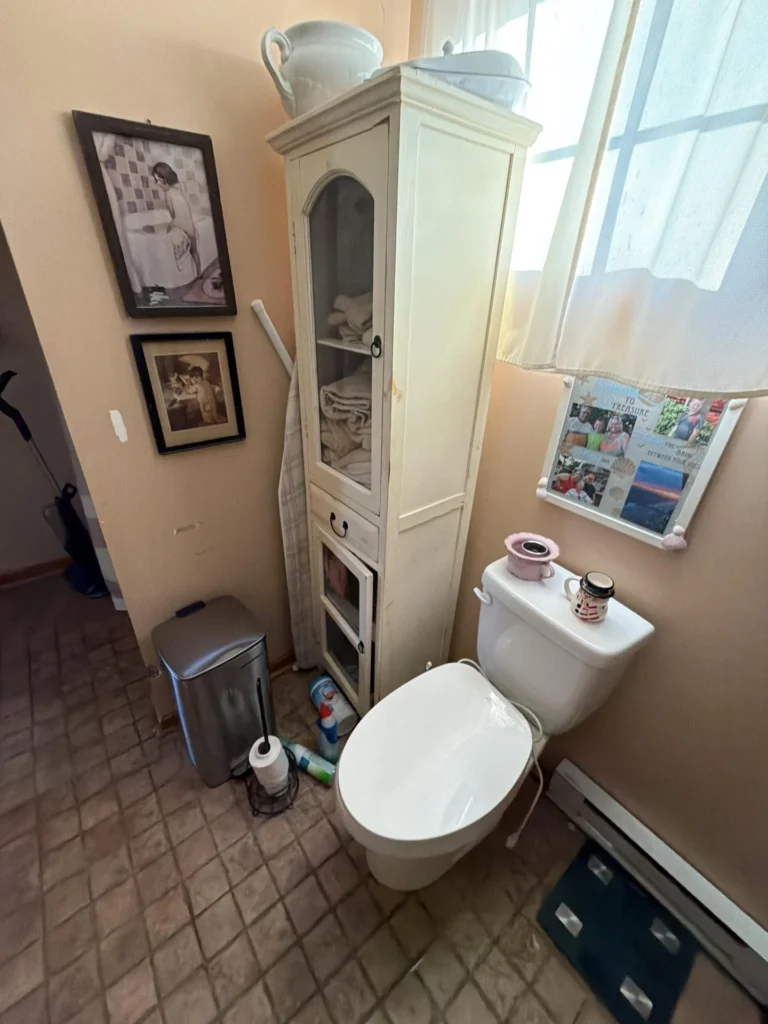 Bathroom with white toilet next to a tall white linen cabinet with glass door, vintage framed wall prints, baseboard heater, and tile floor in a home before remodeling