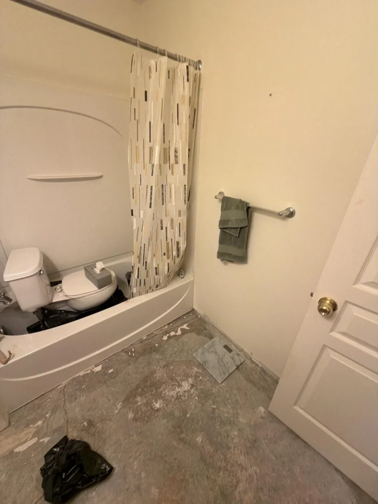Bathroom mid-renovation showing a white soaking tub with patterned shower curtain, bare concrete subfloor with adhesive residue, green sage towel on chrome bar, and white door with brass knob
