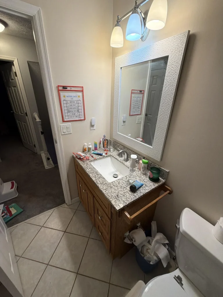 Bathroom with oak vanity, granite countertop, undermount rectangular sink, chrome three-light bar above framed textured mirror, children's morning checklist chart on wall, beige tile floor, open door to carpeted hallway