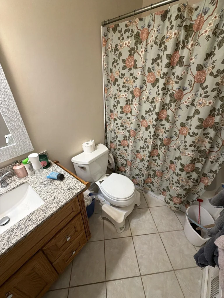 Bathroom with oak wood vanity cabinet, granite countertop with undermount sink, white toilet with step stool, floral botanical shower curtain on chrome rod, beige ceramic tile floor, and laundry bin with towels