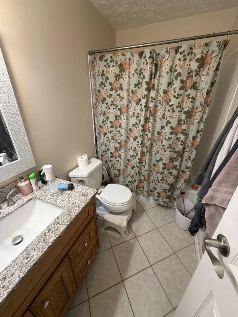 Wide view of bathroom showing oak wood vanity with granite top, white toilet with step stool, floral botanical shower curtain on chrome rod, beige ceramic tile floor, white door with nickel hardware, and partial view of tub and towel