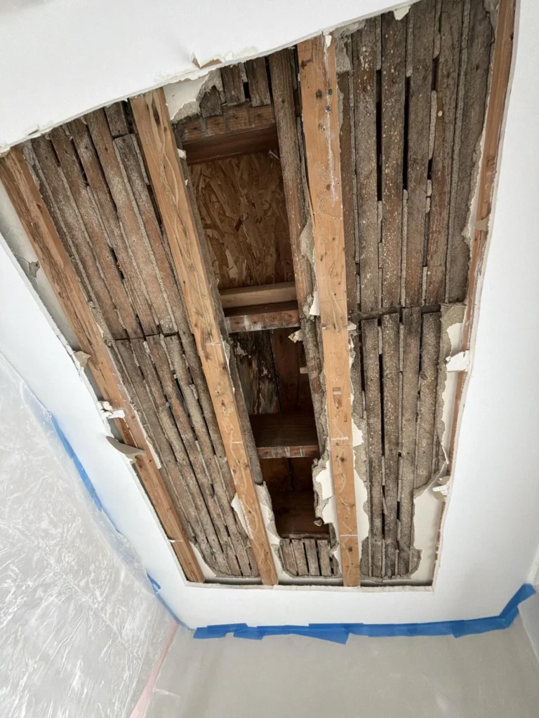 Close-up of a ceiling access opening showing rows of old narrow wooden lath strips and floor joists, with clear plastic sheeting and blue painter's tape around the perimeter protecting surfaces below during remediation