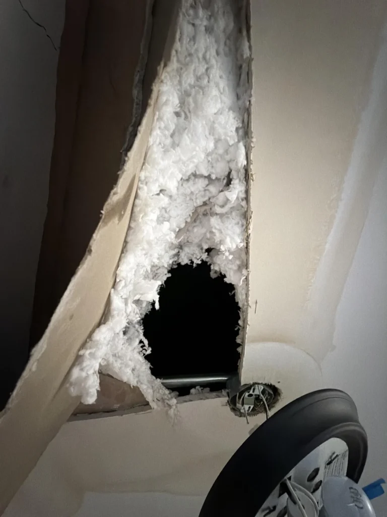 Large jagged hole in ceiling drywall with blown-in white insulation spilling out and dark void above, ceiling light fixture and fan blade visible below