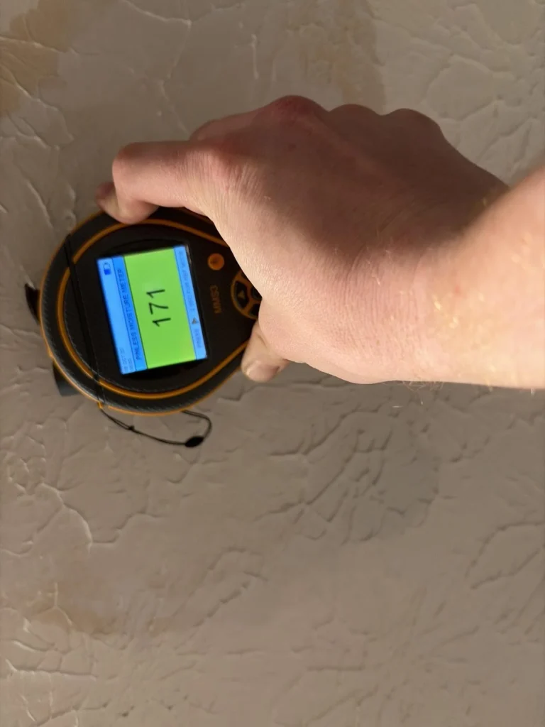 Hand pressing Protimeter MMS3 pinless moisture meter flat against skip-trowel textured ceiling showing reading of 171 on green screen, large brown water stain visible on ceiling surface indicating active moisture from above