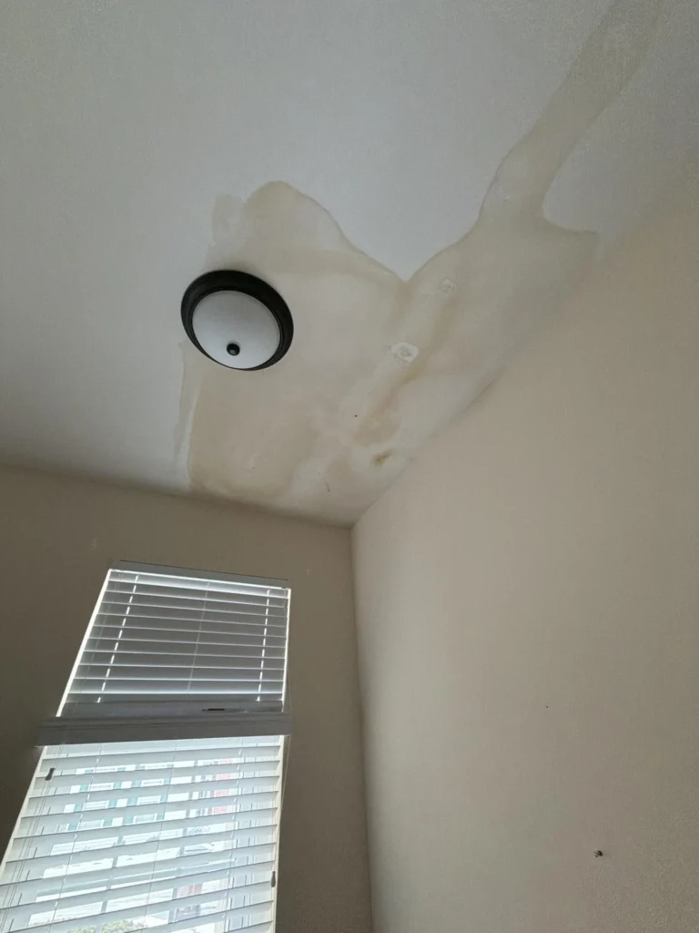 Wide-angle view of room ceiling with large water stain and bulging paint around black dome light fixture, window corner with open blinds, tan walls