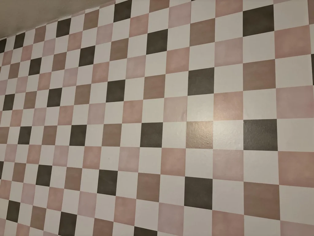 Close-up of a hand-painted checkerboard accent wall in a child's bedroom featuring a multi-tone pattern of white, blush pink, and dark taupe squares in alternating rows