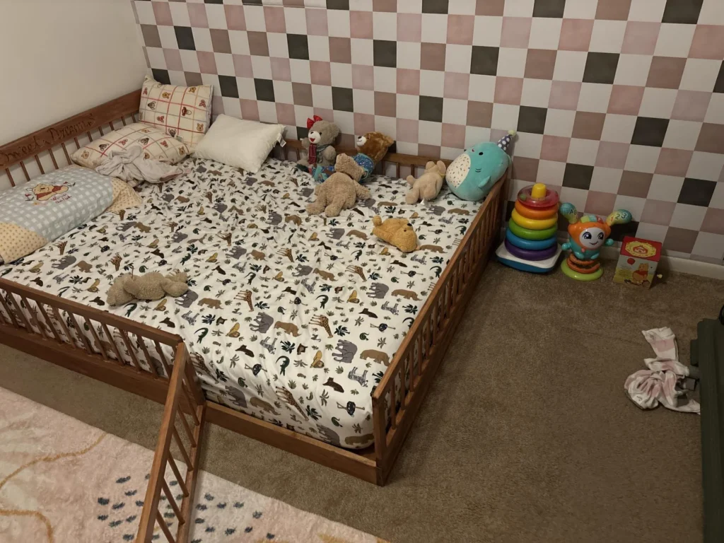 Child's bedroom with wooden floor-bed frame covered in animal-print bedding and multiple stuffed animals, checkerboard accent wall in pink, white, and taupe behind, colorful stacking ring toy and baby toys on carpeted floor beside bed