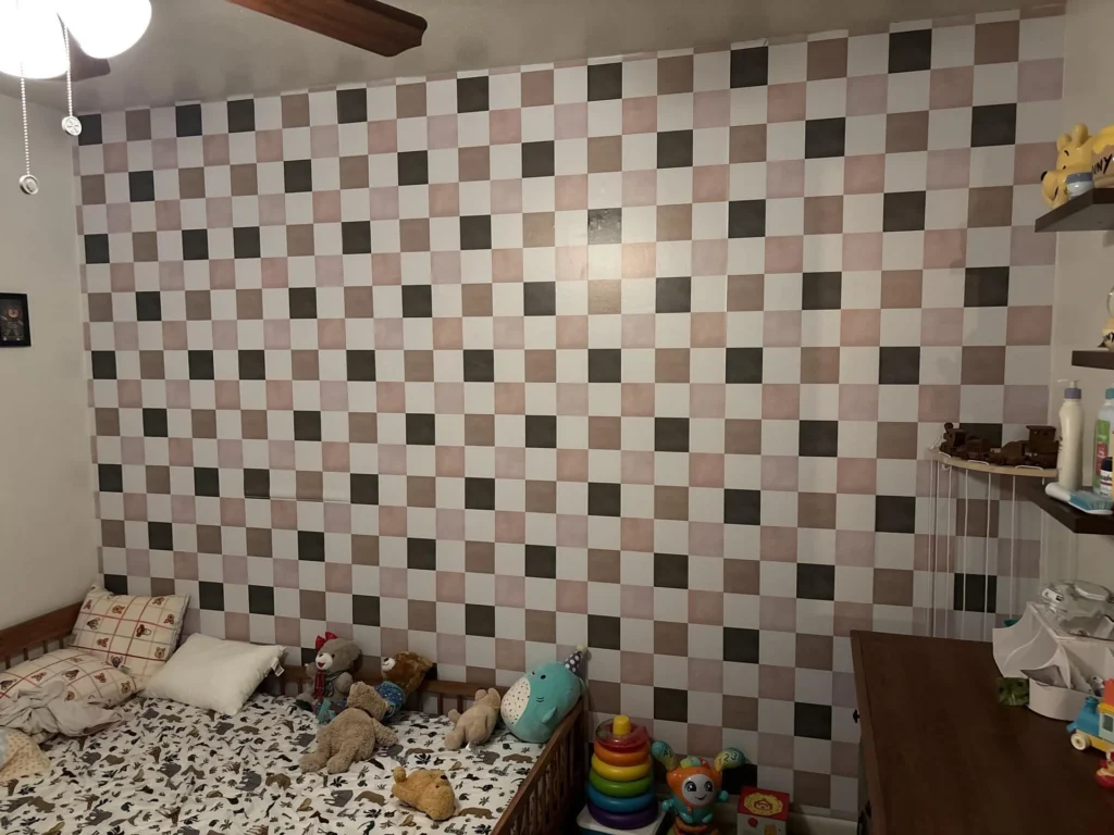 Full wall view of child's bedroom featuring large hand-painted checkerboard accent wall in white, blush pink, and dark taupe, wooden floor-bed with animal-print bedding and stuffed animals in lower left, nursery shelves with baby items on right, ceiling fan with wood blades visible