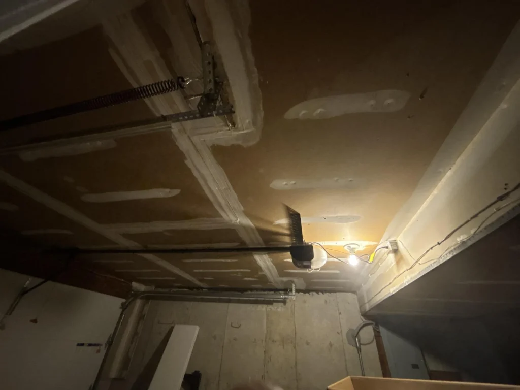 Overhead view of complete garage ceiling with newly installed drywall showing full grid of taped and mudded seams, garage door opener motor and rail centered, torsion spring and track hardware along front wall, exposed electrical wiring on side wall