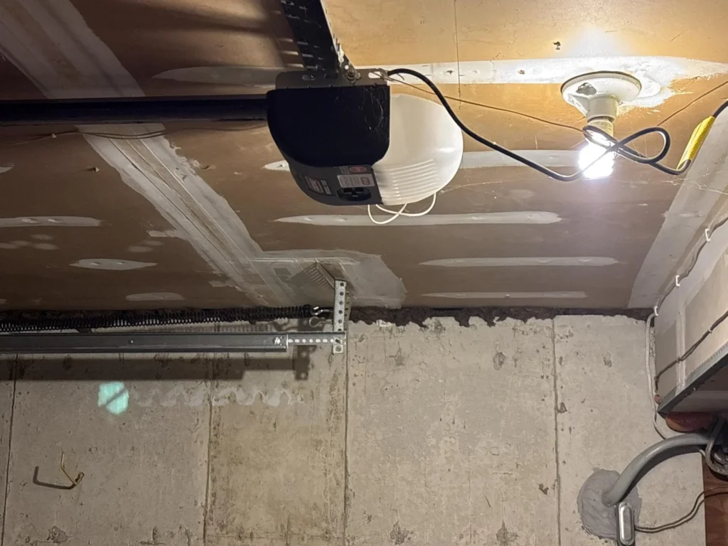 Garage ceiling with newly installed and mudded drywall panels, black garage door opener motor unit mounted to ceiling with white globe light bulb illuminated, wiring visible, concrete block wall with garage door track below