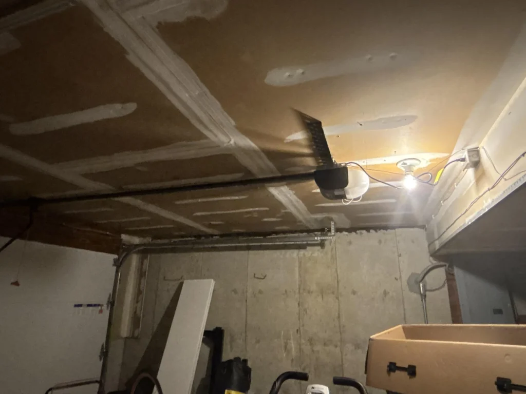 Wide angle view of garage interior showing newly drywalled and mudded ceiling with garage door opener and light, exposed wiring on side wall, cardboard boxes and miscellaneous items on floor, concrete block walls