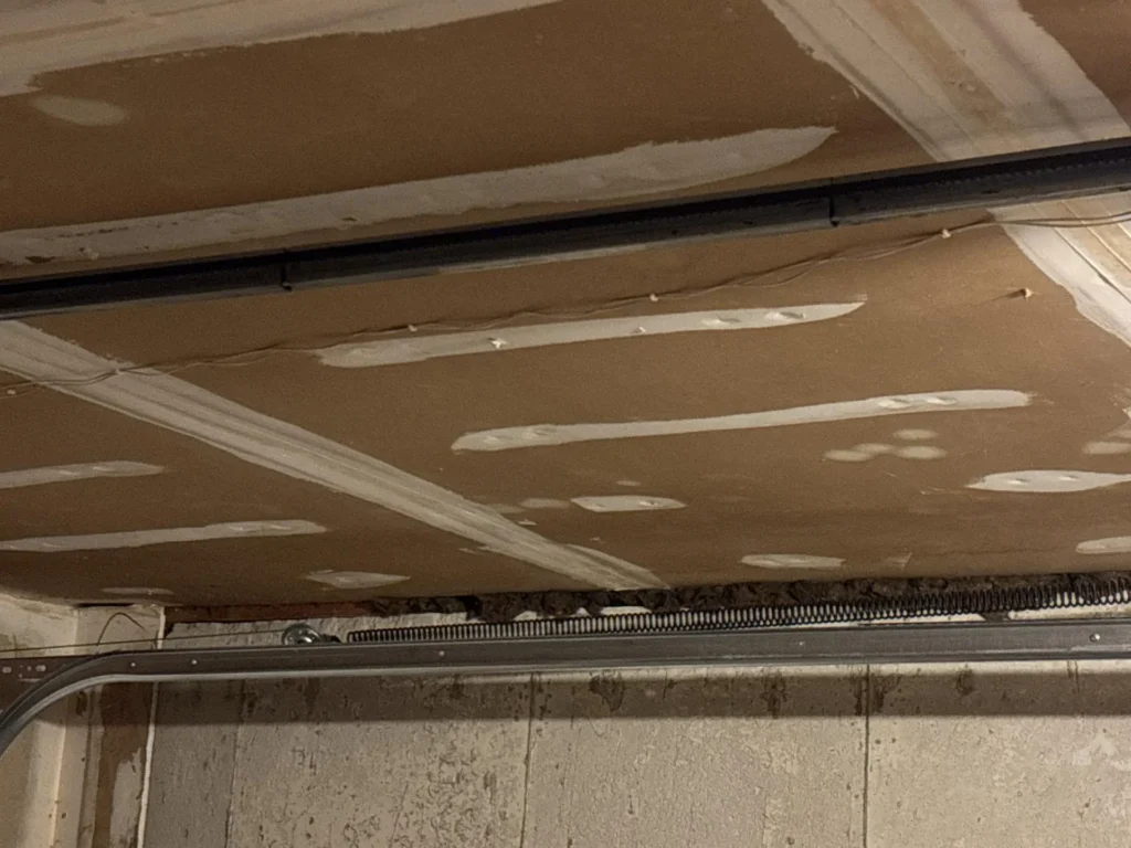 Interior garage ceiling view showing freshly installed drywall with taped and mudded seams, garage door torsion spring and track rail visible along the bottom edge, concrete block wall below