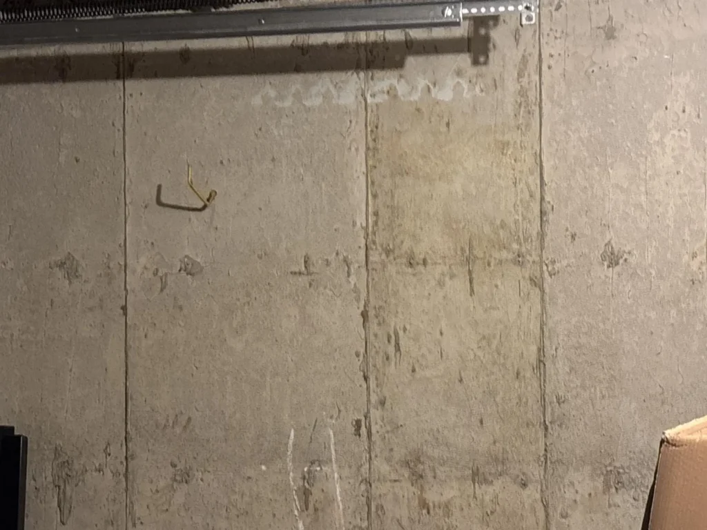 Garage interior concrete block wall showing extensive water staining and efflorescence streaks, garage door horizontal track bracket mounted at top, small bent bracket on wall, cardboard box in lower right corner