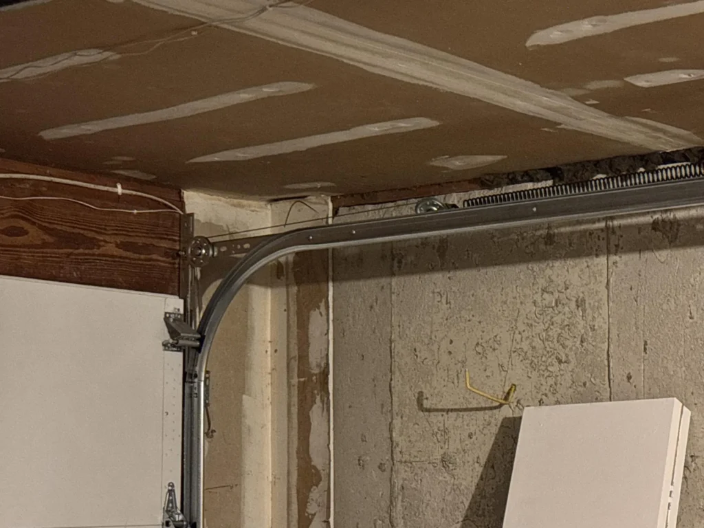 Garage corner showing new drywall ceiling with mudded seams, garage door curved track and torsion spring hardware meeting at wall, wood header above garage door opening, concrete block wall with water damage staining