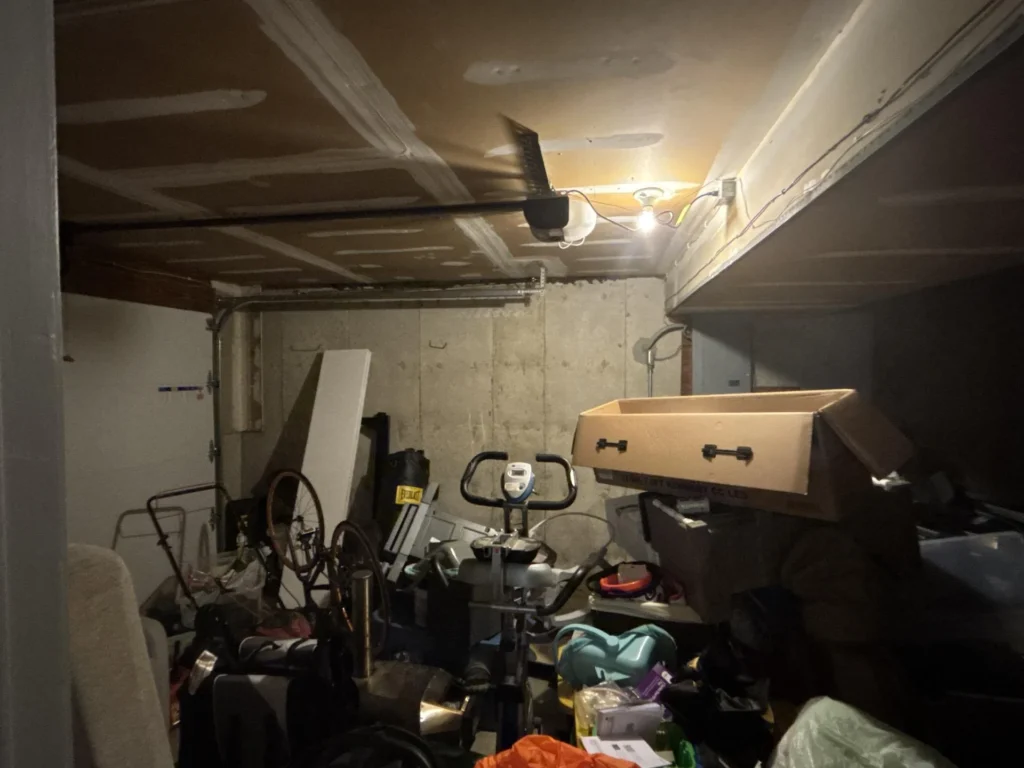 Cluttered garage interior with newly drywalled ceiling and garage door opener, concrete block walls, miscellaneous stored items including bicycles, exercise bike, cardboard boxes, and various household goods on floor