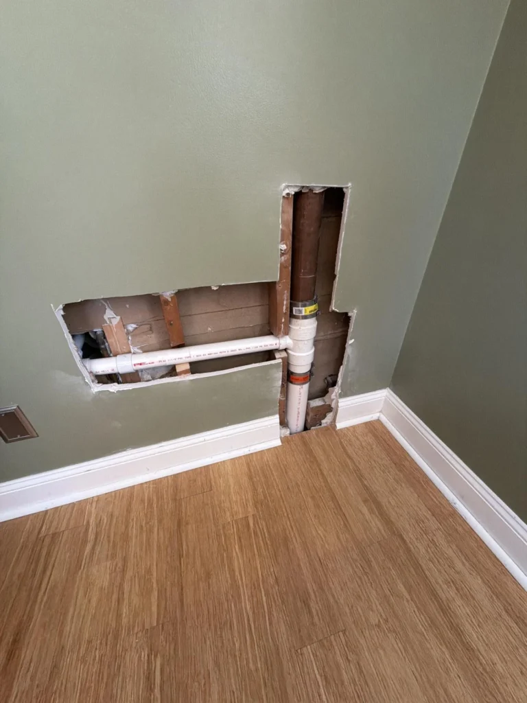 Green-painted bedroom or hallway wall with rectangular drywall cutouts exposing PVC supply pipe and drain pipe connected to copper stack, prior to drywall repair