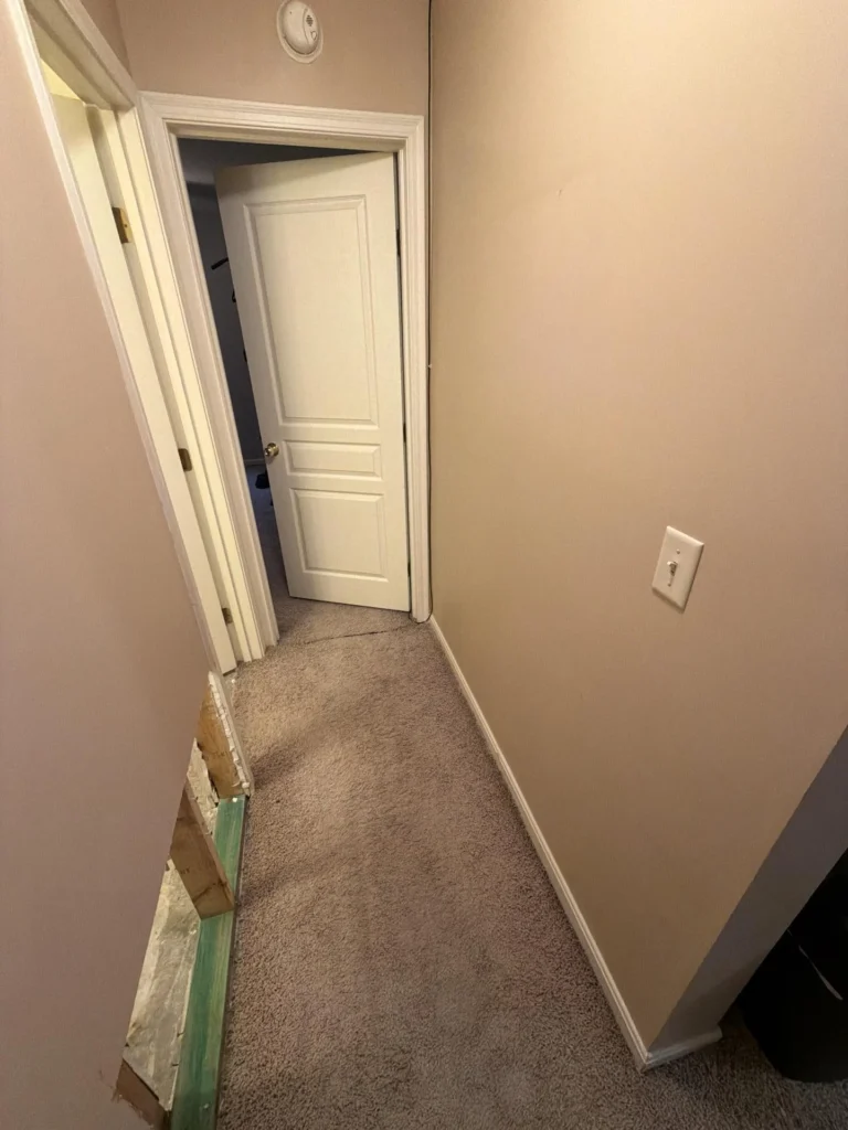 Carpeted hallway with beige walls leading to open bathroom door, drywall cutout at baseboard level on left exposing green-painted wood framing and subfloor, smoke detector on ceiling